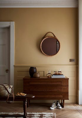 Hallway painted in muted gold shade 'Madeleine' with a wooden sidetable, round mirror and rug.