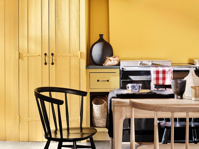 A yellow kitchen  painted in Giallo with a yellow wooden cupboard on the left. There is a cooker, with a gingham red tea towel and a large black vase on the countertop. There is a wooden table infront of the cooker with a black tablecloth and a blue and white cup. There are three chairs at the table, two black ones and a wood one. 