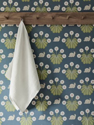 Close-up of snail-printed wallpaper (Burges Snail - Juniper) with brown hooks and a white towel hung on the wall.