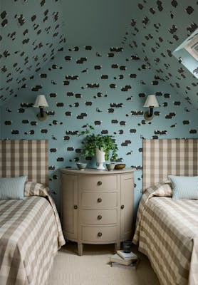 Children's bedroom featuring pale blue dog and sheep print wallpaper (Nip & Lassie - Celestial Blue) with two beds and a bedside drawer in the middle.