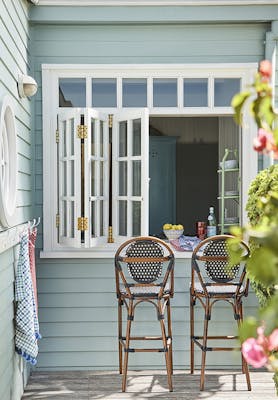 Outside breakfast bar with cladded walls painted in grey blue shade 'Celestial Blue' with two wooden stools under white shutters.