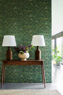 Hallway with green small floral wallpaper 'Millefleur - Garden' and a wooden sidetable with 2 lamps and a vase of flowers.