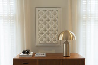 Wooden side table with silver lamp, magazines and a camera in front of a beige Portland Stone wall with white artwork.