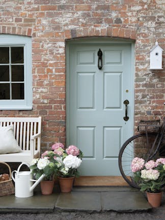 Front door painted in grey blue shade 'Celestial Blue' with a brick surround and a grey bench with flowers.