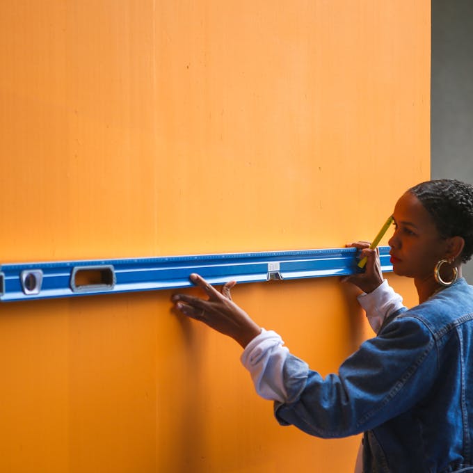 Woman (Lakwena) wearing a denim jacket using a spirit level on a bright orange wall in preparation of painting a mural.