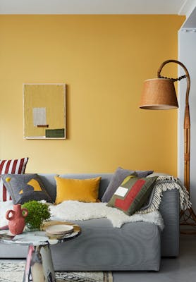 Bright yellow living room painted in Yellow-Pink, with a grey sofa covered in blankets and cushions underneath a lamp.