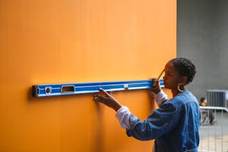 Woman (Lakwena) wearing a denim jacket using a spirit level on a bright orange wall in preparation of painting a mural.