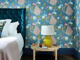 Bedroom featuring blue wallpaper with a vibrant peacock print (Pavona – Sylvie), behind a bed with a green velvet headboard.