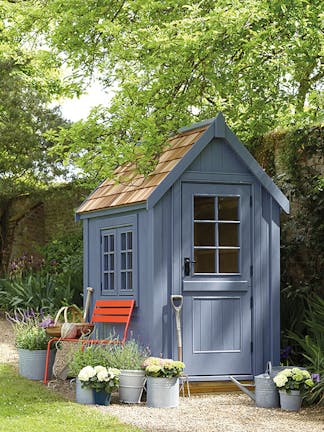 Outdoor shed painted in warm blue 'Juniper Ash' with a bright red chair surrounded by potted plants and trees.