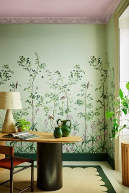 Green floral mural wallpaper featuring birds and butterflies (Bird & Bluebell - Pea Green) with a lamp and plant on a desk.