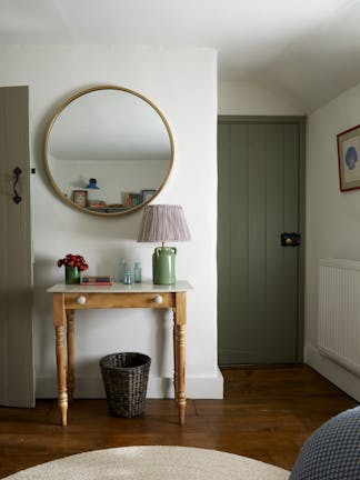 A room featuring two sage green doors, white walls and wood flooring, and a dressing table with a round brass mirror above. 