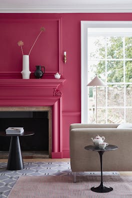 Living space painted in bright pink 'Leather' with a matching fireplace and  contrasting white window frame and an armchair.