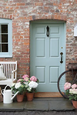 Front door painted in grey blue shade 'Celestial Blue'  with a brick surround and a grey bench with flowers.