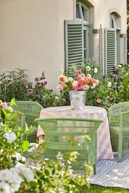 Outside dining area with three bright green garden chairs and a warm neutral wall (Clay) surrounded by flowers and greenery.