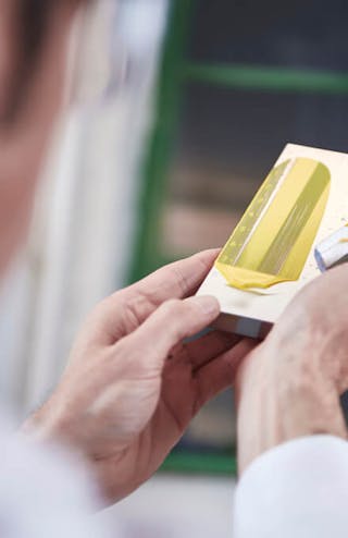 A person in the factory holding a metallic block and spreading yellow Little Greene paint on it with a silver scraper.