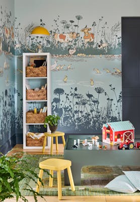 Children's playroom featuring blue mural wallpaper (Riverside Capers - Moo) with storage space to the left alongside yellow stools and toys in the foreground.