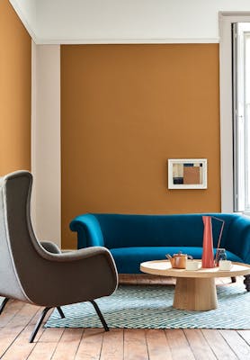 Living space painted in dark orange (Middle Buff) with a contrasting deep blue sofa, grey armchair and wooden table on a rug.