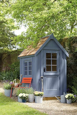 Outdoor shed painted in warm blue 'Juniper Ash' with a bright red chair surrounded by potted plants and trees.