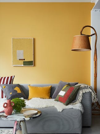 Bright yellow living room painted in Yellow-Pink, with a grey sofa covered in blankets and cushions underneath a lamp.