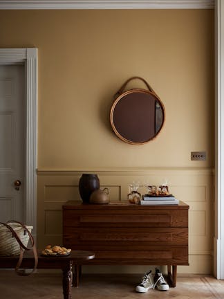 Hallway painted in muted gold shade 'Madeleine' with a wooden sidetable, round mirror and rug.