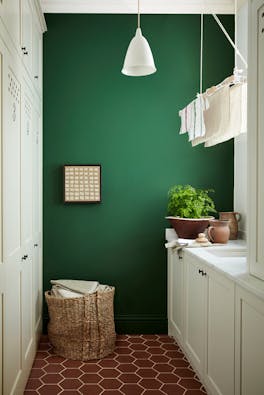 Laundry room painted in dark green shade 'Dark Brunswick' with a laundry basket, white cupboards and tiled floor.