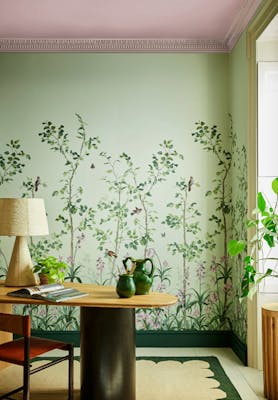 Green floral mural wallpaper featuring birds and butterflies (Bird & Bluebell - Pea Green) with a lamp and plant on a desk.