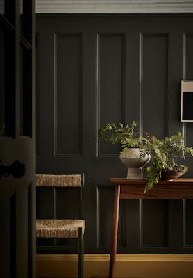 Hallway with dark grey walls (Pompeian Ash), contrasting bright yellow baseboard (Giallo) and side table and wicker chair.