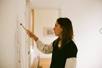 The side profile of a woman using a long paint brush to paint dark outlines on an interior white wall.