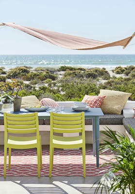 Beachside balcony with a dining table painted in muted blue 'Etruria' and chairs in lime green 'Pale Lime' overlooking the ocean and greenery.