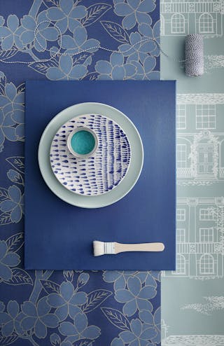 Flat-lay of blue wallpaper samples in different shades and patterns topped with two plates and a blue paint sample pot.