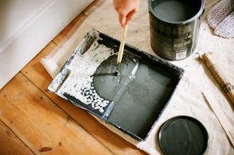 Dark green paint poured onto a paint tray placed next to a paint tip and lid on a partially covered wooden floor.