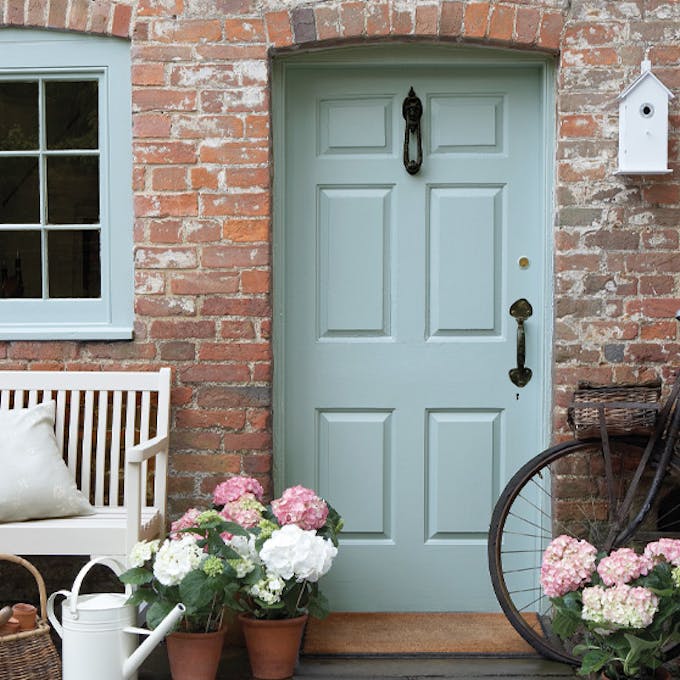 Front door painted in grey blue shade 'Celestial Blue'  with a brick surround and a grey bench with flowers.