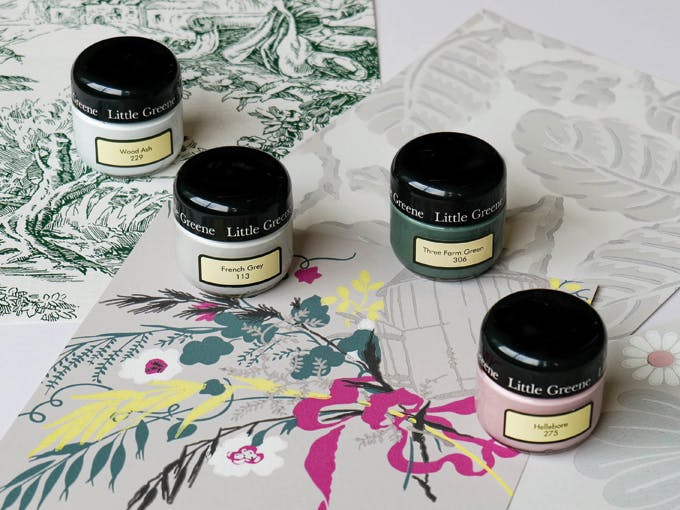 Three Little Greene wallpaper samples, a toile de jouy, a leaf print and a bold flower and bird pattern, four sample pots are placed ontop of the samples in the colours Wood Ash, French Grey, Three Farm Green and Hellebore 