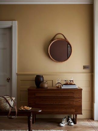 Hallway painted in muted gold shade 'Madeleine' with a wooden sidetable, round mirror and rug.