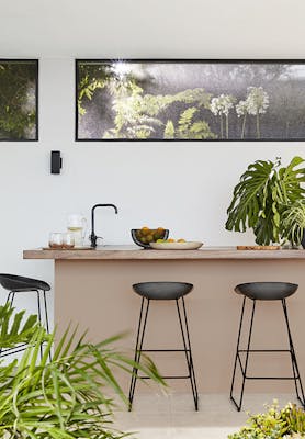 Outside breakfast bar with the back wall painted in off white shade 'Linen Wash' and the island painted in soft pink shade 'Masquerade' with two black stools and plants surrounding.