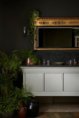 Bathroom with deep brown walls (Elysian Ground) with a white cabinet and a gold mirror above, surrounded by indoor plants.