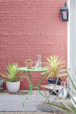 Outside brick wall painted in red shade 'Ashes of Roses' with bright green garden table and pink chair.