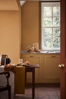 Kitchen space painted in honey shade 'Bombolone' with a paneled window, wooden table and chair.