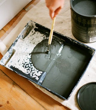 Dark green paint poured onto a paint tray placed next to a paint tip and lid on a partially covered wooden floor.