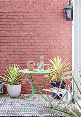 Outside brick wall painted in red shade 'Ashes of Roses' with bright green garden table and pink chair.