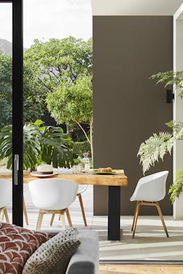 Outside dining space with a wooden table and white chairs surrounded by greenery and a high wall to the right painted in grey-brown shade 'Attic II'.