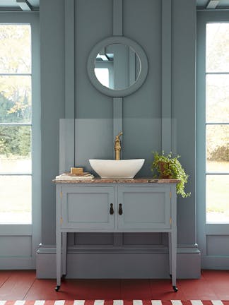 Cloakroom painted in muted blue shade 'Etruria' with a basin and round mirror in the center of two large windows.