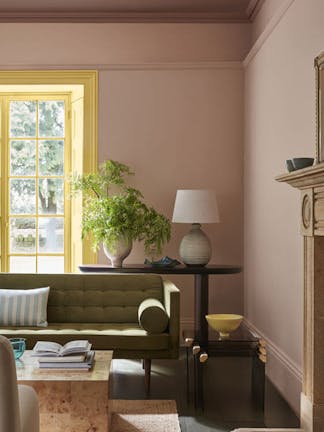 Living space painted in muted pink shade 'Masquerade' with a contrasting yellow 'Light Gold' window frame and an olive green sofa in the foreground.